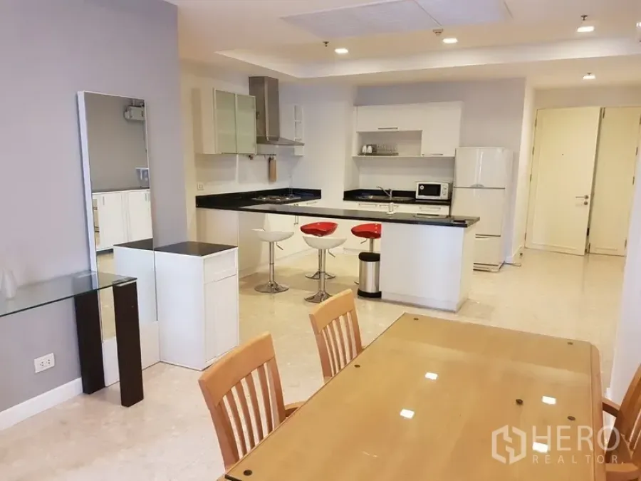 Condominium for rent in Khlong Toei, Bangkok - Open-plan kitchen with white cabinetry, black stone countertops, island breakfast bar with red stools, and adjacent dining area.