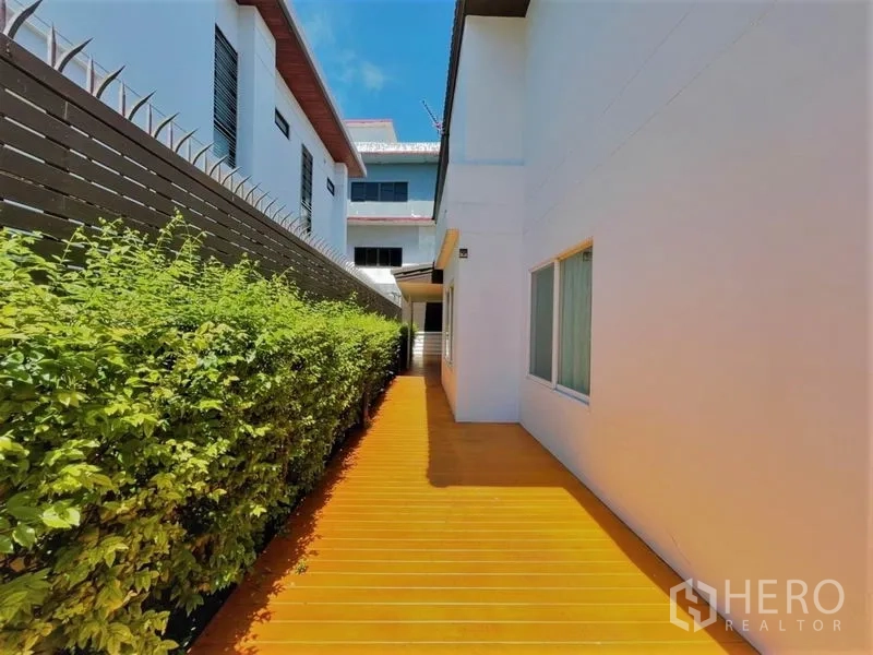 Detached House for rent in Suan Luang, Bangkok - Sunlit walkway along wooden deck bordered by privacy fence and manicured hedge.