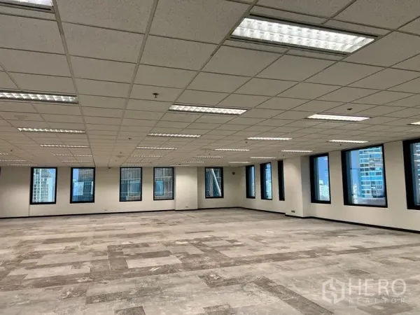 Office Space for rent in Khlong Toei, Bangkok - Large office interior with partially tiled flooring, recessed fluorescent lighting, and skyline views through black‑trimmed windows.