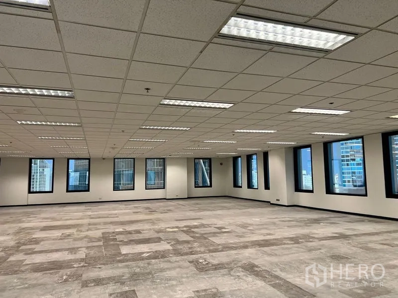 Office Space for rent in Khlong Toei, Bangkok - Large office interior with partially tiled flooring, recessed fluorescent lighting, and skyline views through black‑trimmed windows.