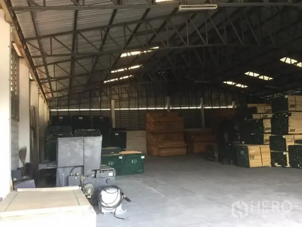 Warehouse for sale in Sai Noi, Nonthaburi - Warehouse interior showing steel truss roof, daylight panels, and stacked materials on a concrete floor.