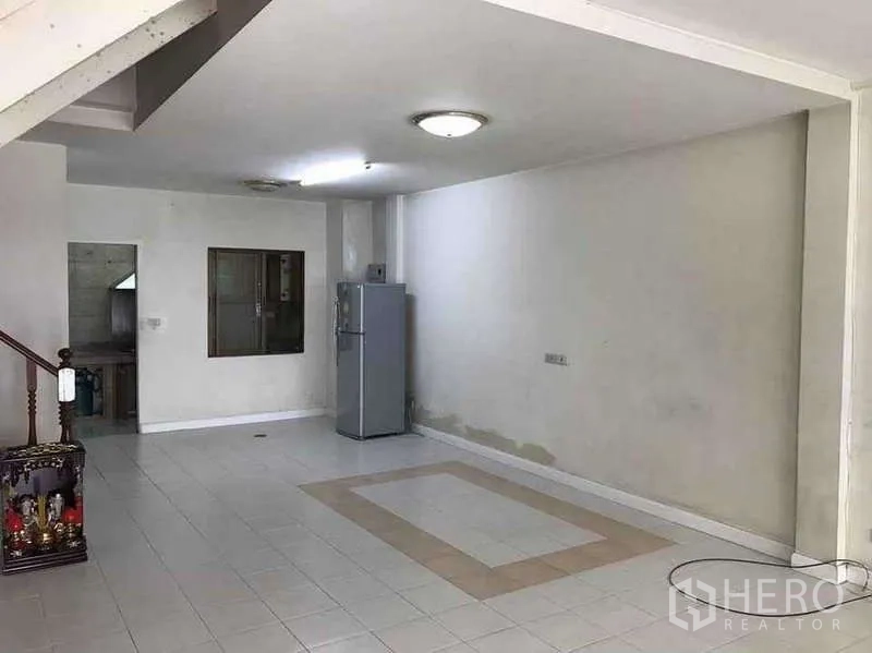 Townhouse for rent in Suan Luang, Bangkok - Open-plan living area with white tiled floor, ceiling lights and a refrigerator leading toward the kitchen.