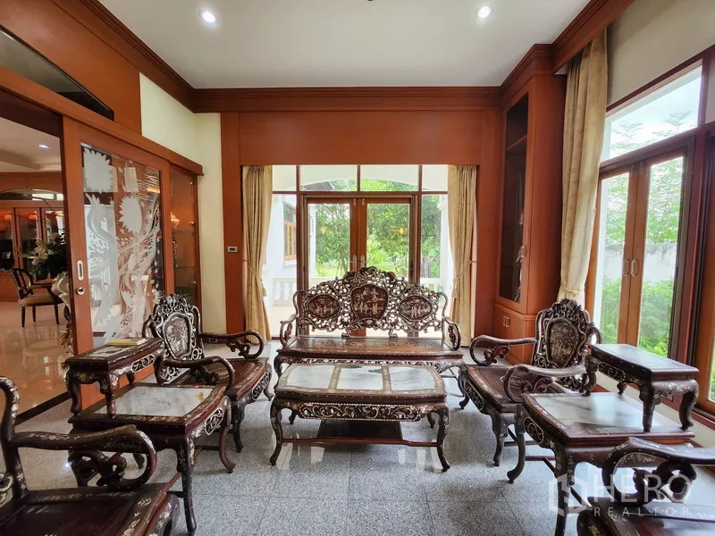 Detached House for rent in Prawet, Bangkok - Cozy sitting room with ornate carved wooden furniture and glass doors opening to green garden views.