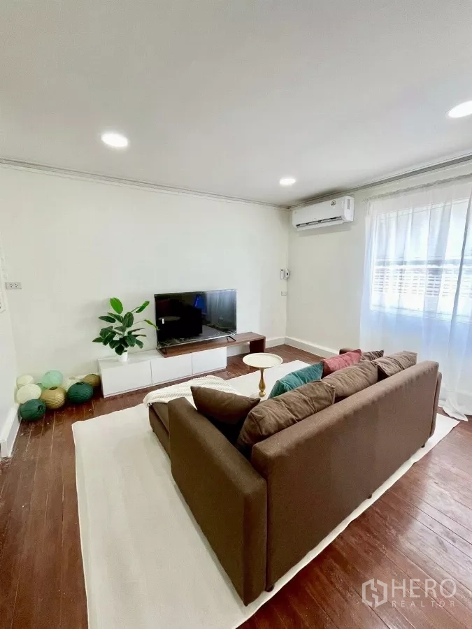 Detached House for rent in Chatuchak, Bangkok - Bright living room with brown sectional sofa, TV console, wood floors and sheer curtains.