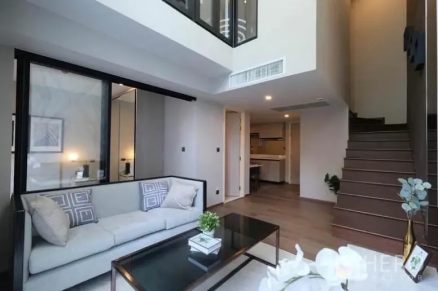 Condominium for rent in Pathum Wan, Bangkok - Double-height living room with staircase, sofa and an open kitchen in a modern condominium.