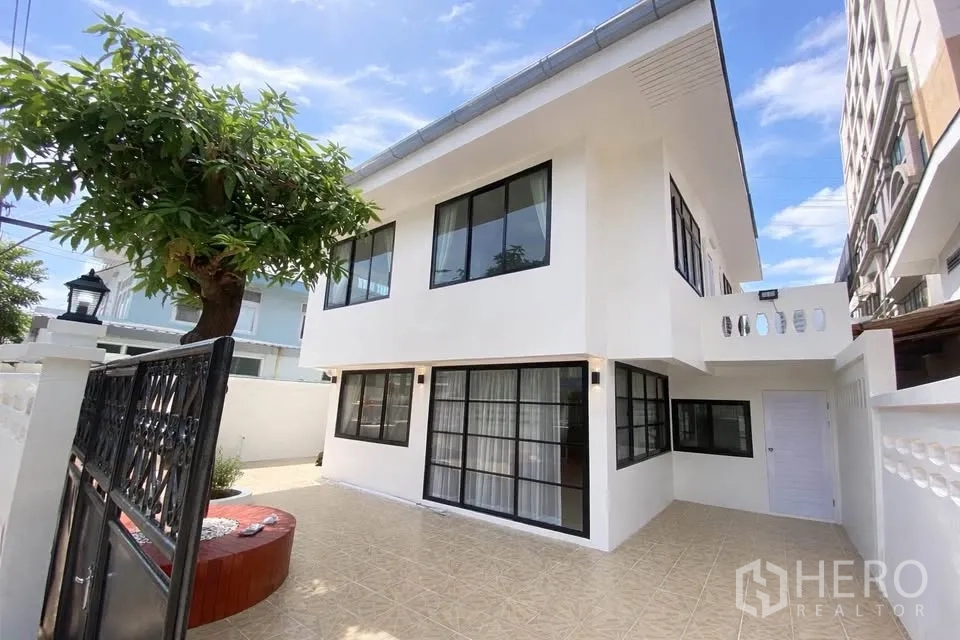 Detached House for rent in Huai Khwang, Bangkok - Exterior of two-storey white detached house with tiled courtyard and tree behind gate.