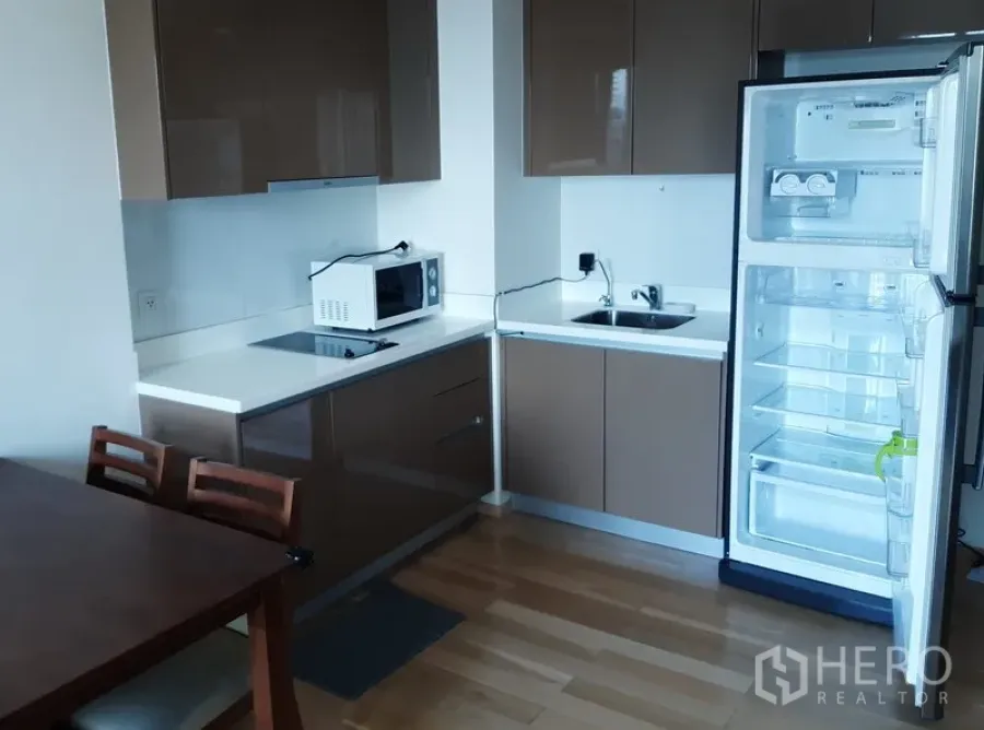 Condominium for rent in Khlong Toei, Bangkok - Modern compact kitchen with glossy cabinets, cooktop, microwave, sink, and open refrigerator on wood flooring.