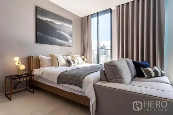 Condominium for rent in Pathum Wan, Bangkok - Serene bedroom with king bed, bedside lamps and large artwork next to a tall window overlooking the skyline.
