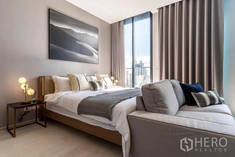 Condominium for rent in Pathum Wan, Bangkok - Serene bedroom with king bed, bedside lamps and large artwork next to a tall window overlooking the skyline.