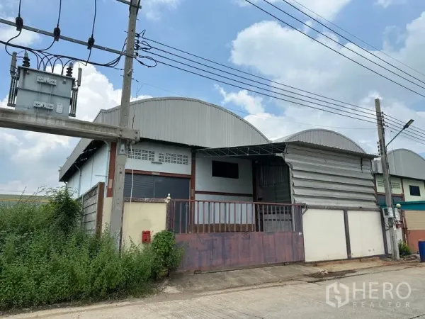 Warehouse for rent in Pak Kret, Nonthaburi - Angled view of metal-clad warehouse row with gated loading area, utility pole and transformer along a concrete road in Nonthaburi.
