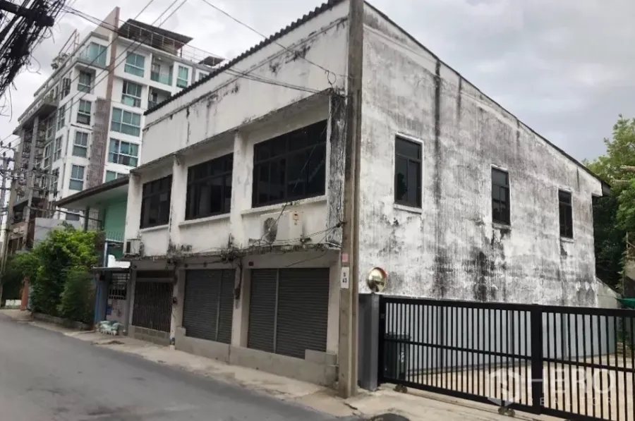 Land for sale in Phra Khanong, Bangkok - Corner view of a two-storey concrete building and gated land on a quiet Phra Khanong lane with condominiums in the background.