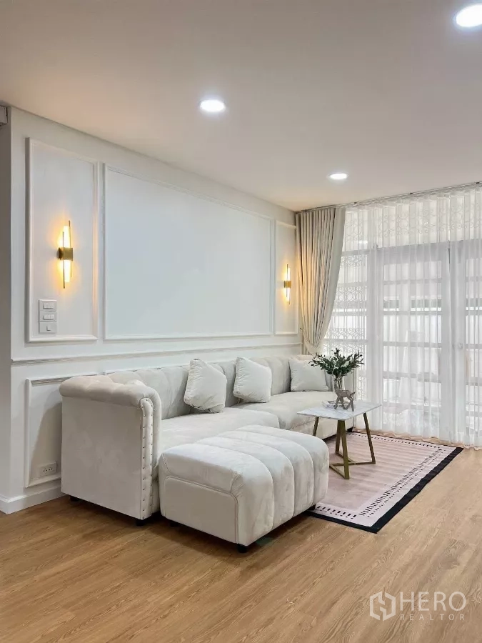 Detached House for rent in Huai Khwang, Bangkok - Corner view of living room highlighting tufted sectional, area rug and warm wall lights.