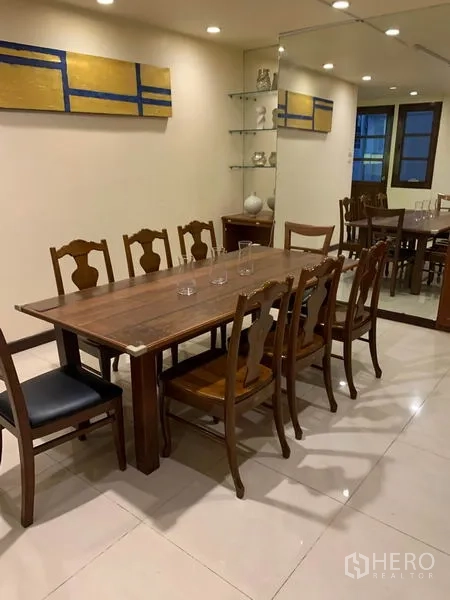 Townhouse for rent or sale in Phra Khanong, Bangkok - Long wooden dining table with chairs beside a mirrored wall and glass shelves.