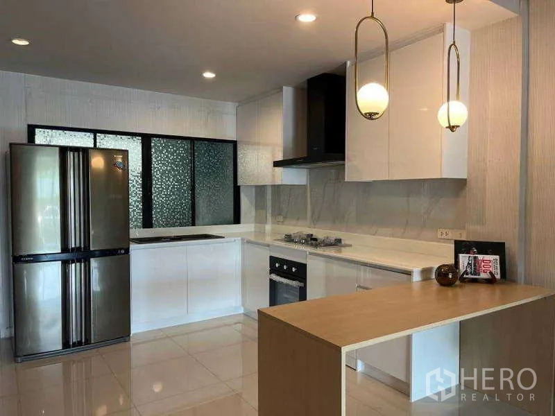 Townhouse for rent in Khlong Toei, Bangkok - L-shaped kitchen with double-door fridge, glossy cabinets, and peninsula lighting.
