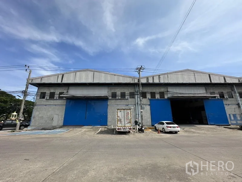 Warehouse for rent in Bang Phli, Samut Prakan - Front elevation showing multiple blue steel doors and parking spaces in front of the warehouse.