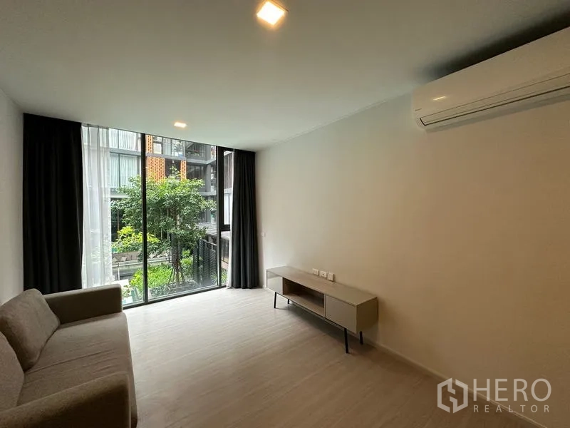 Condominium for rent in Khlong Toei, Bangkok - Minimalist lounge with sofa, TV console, air conditioning, and sliding glass doors overlooking the garden and pool.