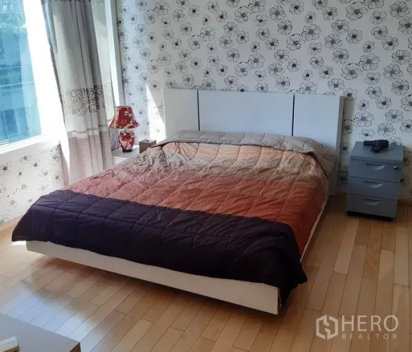 Condominium for rent in Khlong Toei, Bangkok - Bright bedroom with large window, queen bed, bedside tables and floral feature wall on light wooden floors.