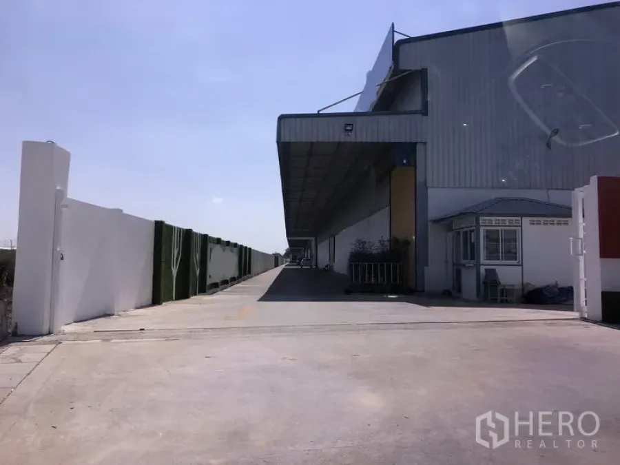 Warehouse for rent in Lat Krabang, Bangkok - Exterior of a modern Lat Krabang warehouse with a long covered loading bay, wide concrete driveway, perimeter wall, and a small guard booth under a clear sky.