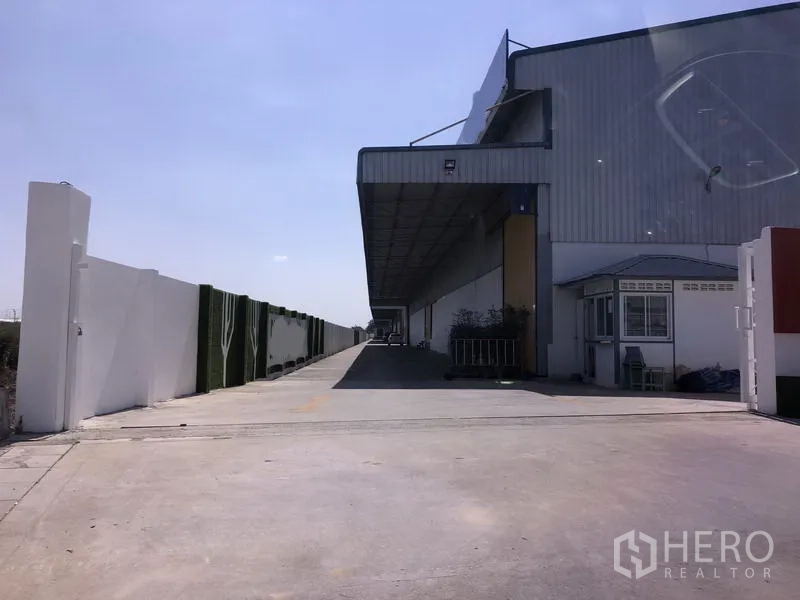 Warehouse for rent in Lat Krabang, Bangkok - Exterior of a modern Lat Krabang warehouse with a long covered loading bay, wide concrete driveway, perimeter wall, and a small guard booth under a clear sky.