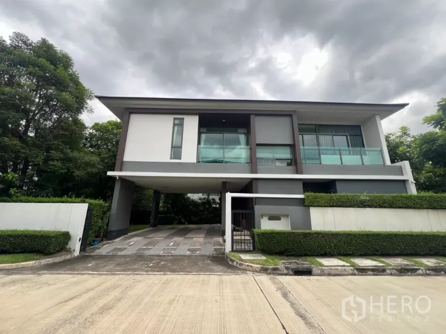 Detached House for rent in Bang Kapi, Bangkok - Modern two-storey corner house with glass balcony, carport and manicured hedges under cloudy sky.