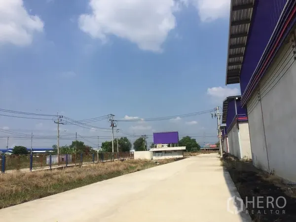 Warehouse for rent in Sai Noi, Nonthaburi - Wide private concrete access road beside warehouse buildings with perimeter fencing and overhead power lines.