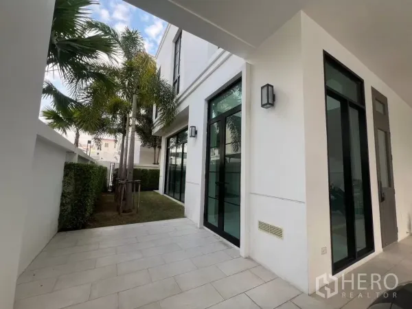 Detached House for rent in Lat Phrao, Bangkok - Side patio and small lawn with palm trees, hedges and black-framed glass doors of the white detached house.