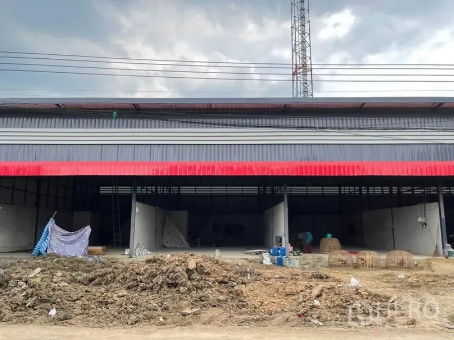 Warehouse for rent in Pathum Thani - Street-facing exterior of a metal-clad warehouse with wide open bays and a dirt work area in front.