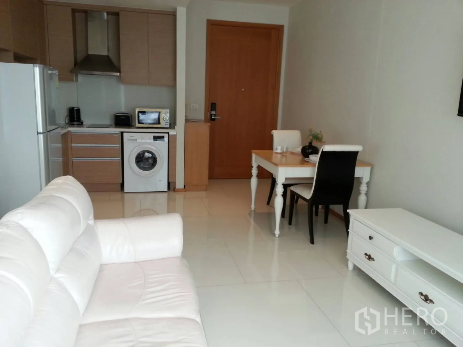 Condominium for rent in Khlong Toei, Bangkok - Open-plan living room with white sofa, compact kitchen with washing machine, and dining table in a Sukhumvit condo.