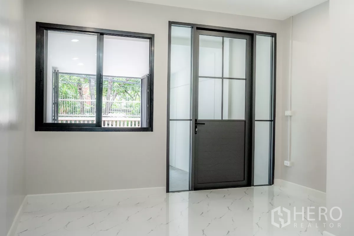 Townhouse for sale in Watthana, Bangkok - Ground-floor multipurpose room with a window and black-framed glass entry door.
