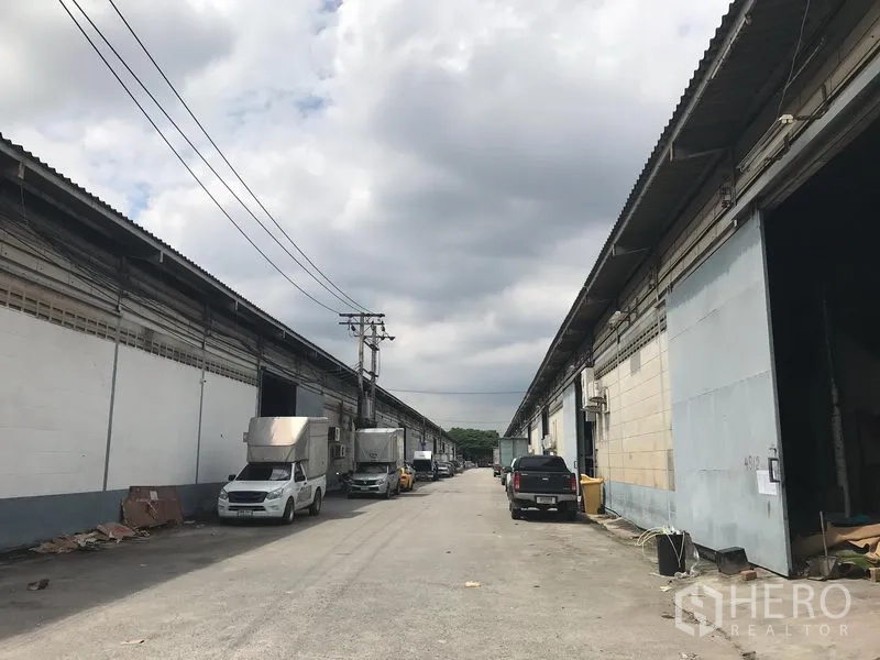 Warehouse for rent in Bang Phli, Samut Prakan - Row of warehouses with wide access road and parked trucks under a cloudy sky.