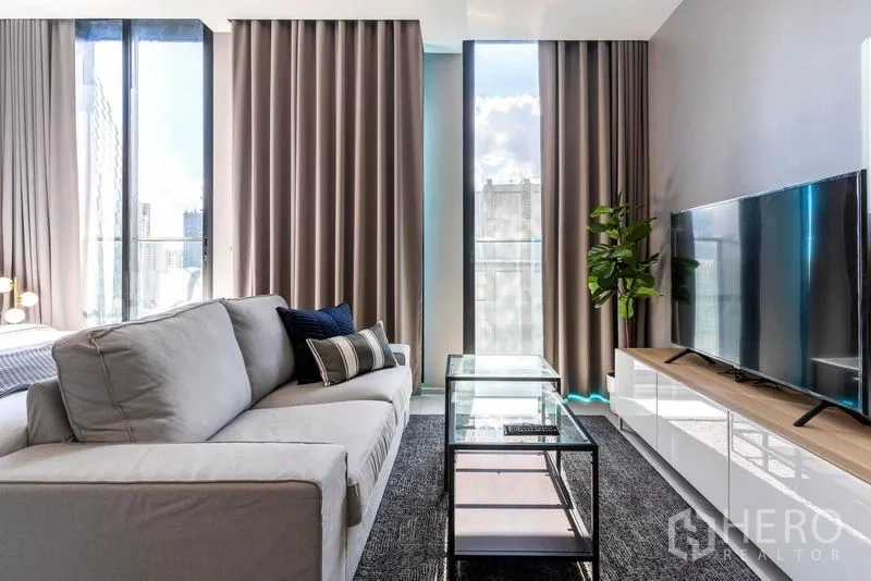 Condominium for rent in Pathum Wan, Bangkok - Bright living area with grey sofa, glass coffee table, TV console and floor-to-ceiling windows offering a city view.