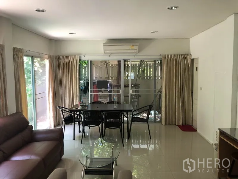 Townhouse for rent or sale in Suan Luang, Bangkok - Bright living–dining area of a Noble Cube Suan Luang townhouse with leather sofa, glass table and sliding doors to a green balcony.