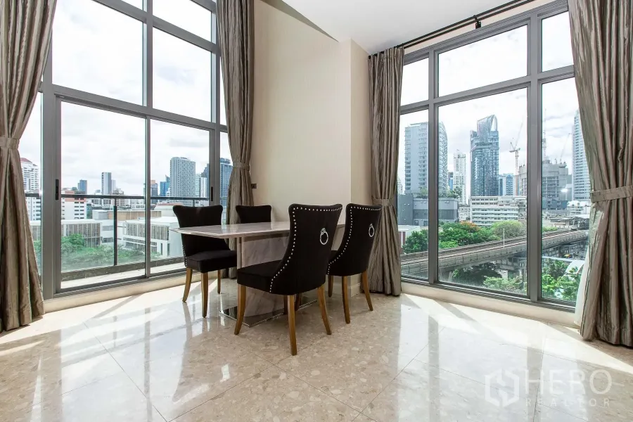 Condominium for rent in Khlong Toei, Bangkok - Corner dining area with marble table and four chairs beside floor-to-ceiling windows overlooking the Bangkok skyline and BTS track.