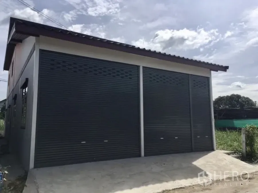Warehouse for rent in Lam Luk Ka, Pathum Thani - Modern warehouse exterior with dual steel roller shutters and a concrete driveway under a pitched roof.