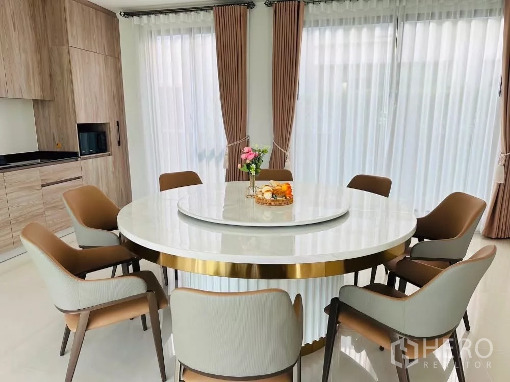 Detached House for rent or sale in Bang Kapi, Bangkok - Dining room with round marble table for eight beside large windows and built-in kitchen.