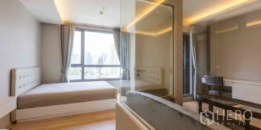 Condominium for rent or sale in Watthana, Bangkok - Bedroom with floor-to-ceiling window, city skyline view, and a glass-walled bathroom with bathtub.