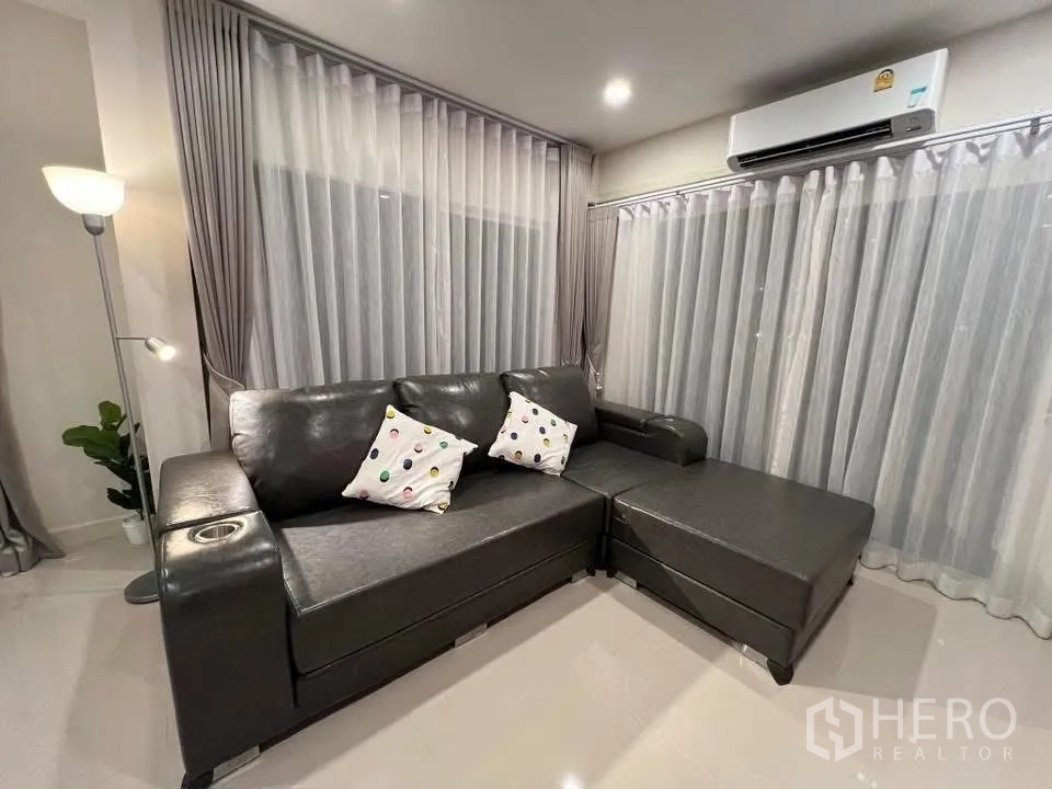 Detached House for rent in Prawet, Bangkok - Living room with L-shaped leather sofa, floor lamp, AC and sheer curtains.
