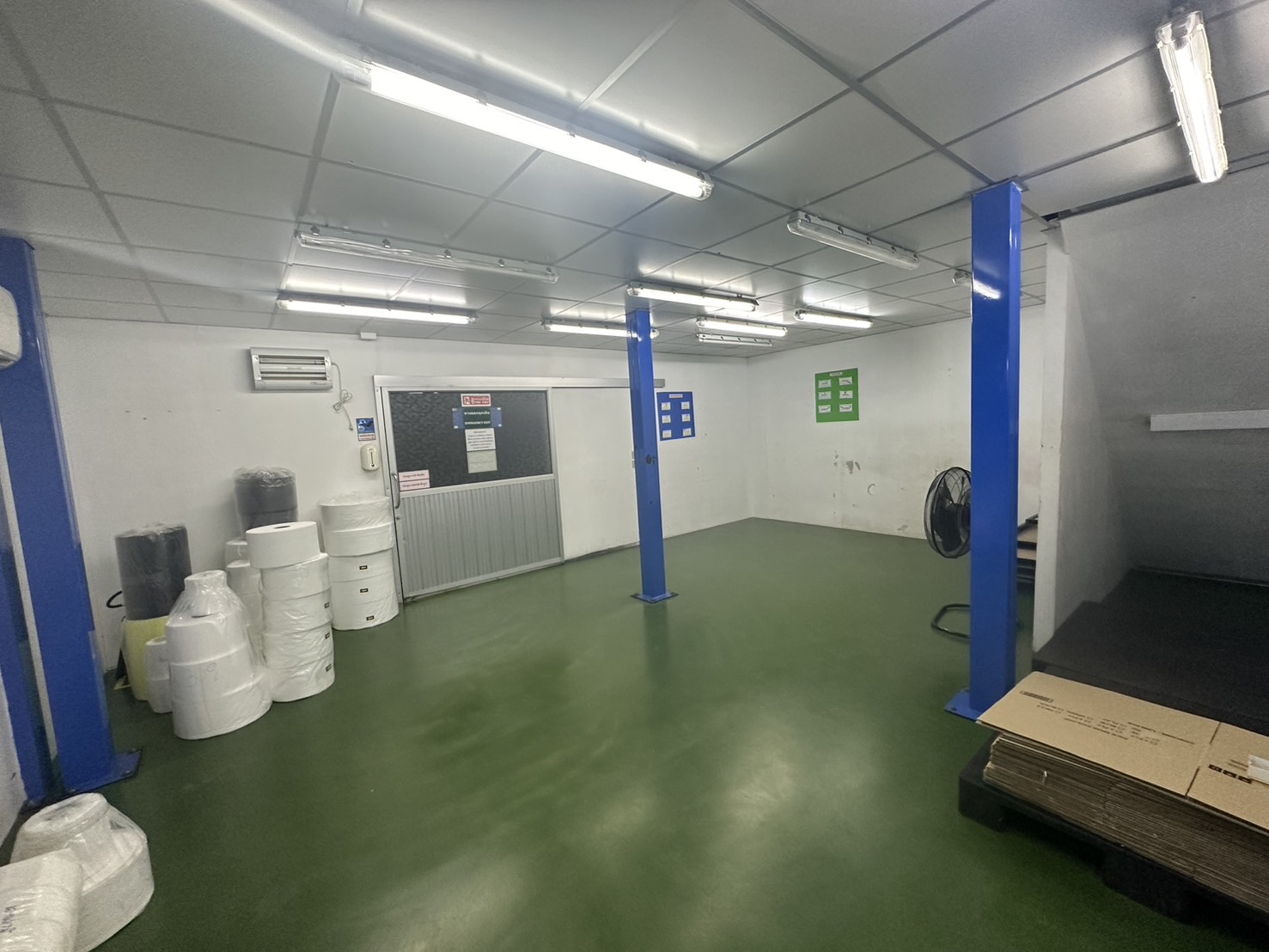 Warehouse for rent or sale in Lam Luk Ka, Pathum Thani - Open workshop zone with blue steel pillars, AC units and rolls of packing material on a green floor.