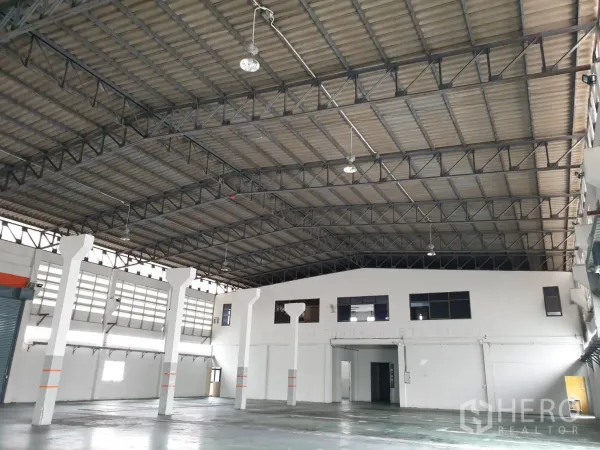 Factory for rent in Bang Pakong, Chachoengsao - Wide warehouse interior featuring a high steel-truss roof, hanging lights and white support columns.