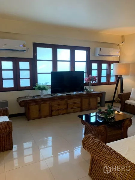 Townhouse for rent or sale in Phra Khanong, Bangkok - Air-conditioned living room with rattan sofa set, TV console and a wall of windows.