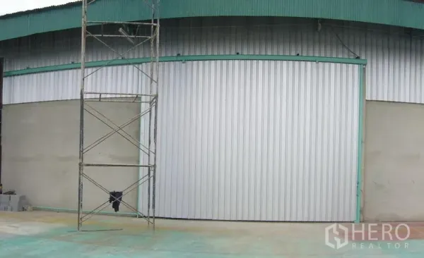 Warehouse for rent in Mueang Pathum Thani, Pathum Thani - Exterior of an industrial warehouse with a wide metal sliding door, teal roof trim, and a concrete apron.