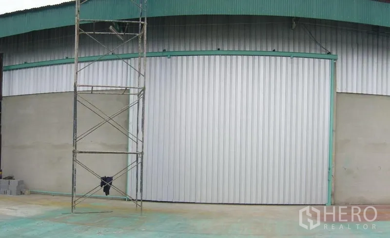 Warehouse for rent in Mueang Pathum Thani, Pathum Thani - Exterior of an industrial warehouse with a wide metal sliding door, teal roof trim, and a concrete apron.