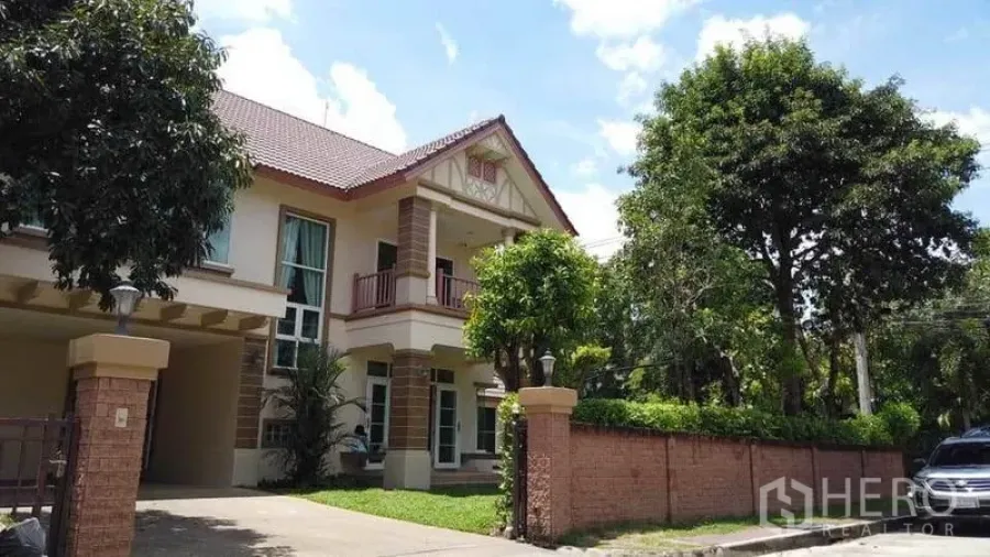 Detached House for rent or sale in Pak Kret, Nonthaburi - Street view of a two-storey house behind a brick fence with driveway and large trees.