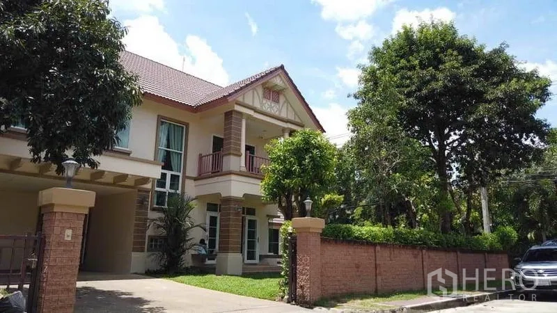 Detached House for rent or sale in Pak Kret, Nonthaburi - Street view of a two-storey house behind a brick fence with driveway and large trees.