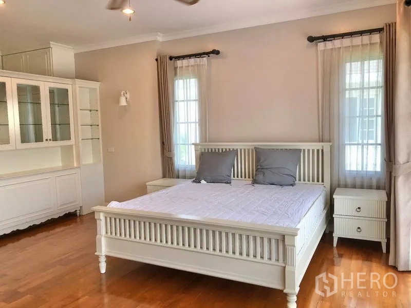 Detached House for rent in Mueang Samut Prakan, Samut Prakan - Master bedroom with wooden floors, white bed frame, bedside tables, built‑in cabinetry and tall windows.