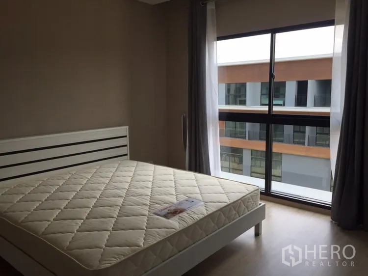 Townhouse for rent in Suan Luang, Bangkok - Bedroom with a large window, neutral walls, and a white bed frame overlooking nearby buildings.