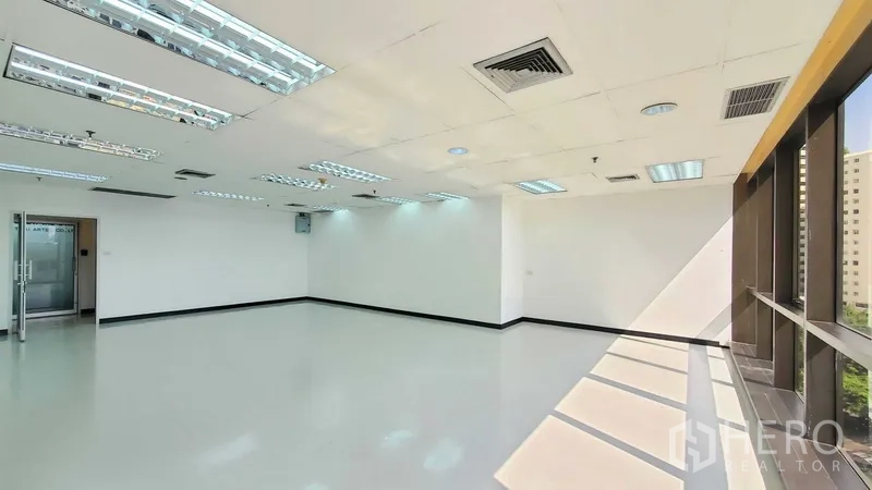 Office Space for rent in Watthana, Bangkok - Corner office interior with window wall, glossy flooring, glass entry door, and ceiling air vents illuminated by natural light.