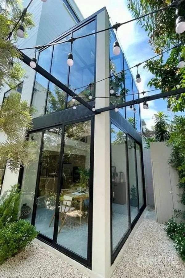Townhouse for sale in Khlong Toei, Bangkok - Modern corner facade with floor-to-ceiling windows and string lights in the townhouse garden.