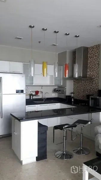 Condominium for rent in Khlong Luang, Pathum Thani - Modern open-plan kitchen with white cabinets, black granite island, colorful pendant lights and two bar stools.