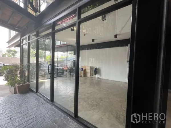 Retail Space for rent - Wide glass façade under canopy with bright interior; potted plants and parked cars outside.